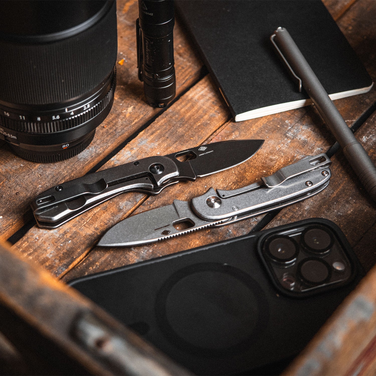 Two pocket knives on a wooden surface with a camera lens and other items.