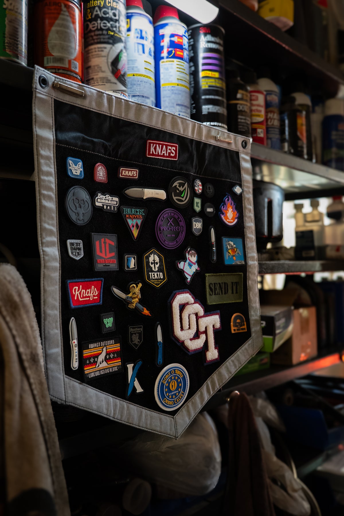 A black canvas patch pocket organizer with a silver-gray stitched border, filled with various automotive and motorcycle decals, stickers, and patches. The organizer is hanging on a shelf next to spray cans.
